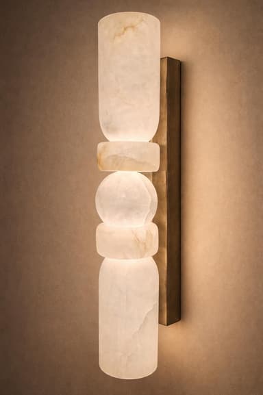 A vertical wall-mounted lighting fixture featuring stacked, irregular, organic-shaped elements made from translucent white and cream onyx. These stone sections are illuminated from within, diffusing a soft glow. The onyx elements are mounted onto a rectangular, brushed brass backplate that runs vertically along the fixture's length. — 3D model