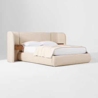 A platform bed with an integrated upholstered headboard that extends to the sides, creating wing-like panels. Two small, wooden nightstands are mounted on the wing panels, one on each side. The bed frame and headboard are upholstered in a textured, light-colored fabric. The mattress is covered with white linens and a light beige throw blanket. — 3D model