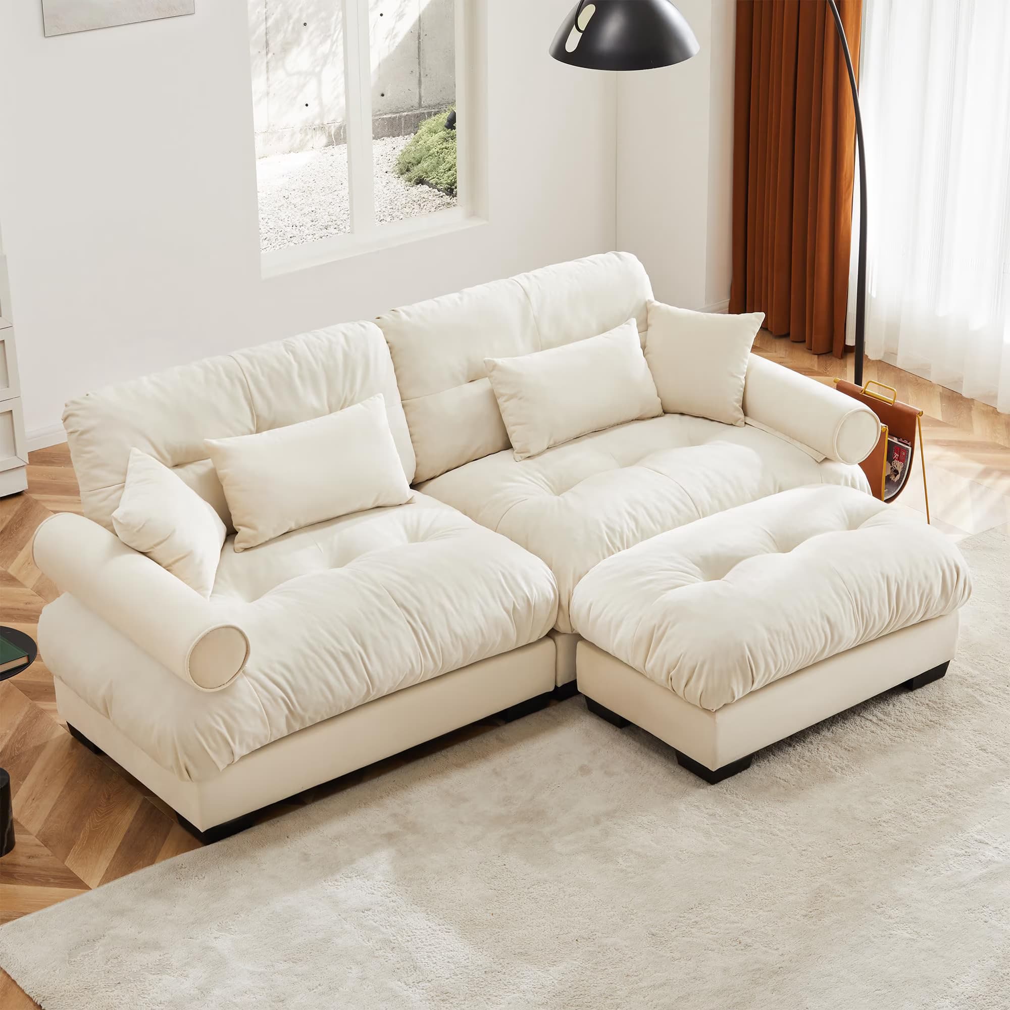 This image features a contemporary cream-colored modular sofa with a chaise component. The sofa is upholstered in a soft, plush fabric and features deep, tufted cushions on both the seat and backrests. It includes several throw pillows and cylindrical bolster pillows integrated into the armrests. The design is low-profile with dark, square feet, suggesting a focus on comfort and modern aesthetics. — 3D model