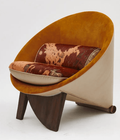 An distinctive lounge chair featuring a deep, curved shell form. The interior of the shell is upholstered in a warm, amber-toned velvet or suede-like fabric, providing a cocoon-like seating experience. The seat cushion and a bolster pillow are upholstered in a bold brown and white cowhide pattern, contrasting with the outer beige or cream-colored leather/vinyl covering the chair's exterior. The chair is supported by two thick, angled dark wooden legs, creating a stable and visually striking base. A brass-colored metallic element appears at the base of the rear, adding a subtle detail. — 3D model