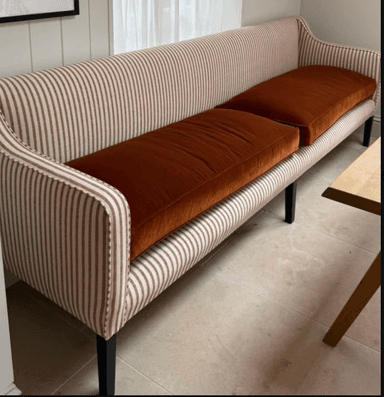 A long, upholstered settee or banquette featuring a high, slightly curved back and subtle, integrated armrests. It is upholstered in a vertical striped fabric in beige and brown, complemented by two solid orange seat cushions. The piece is accented with decorative nailhead trim along the edges and supported by four dark, tapered wooden legs. — 3D model