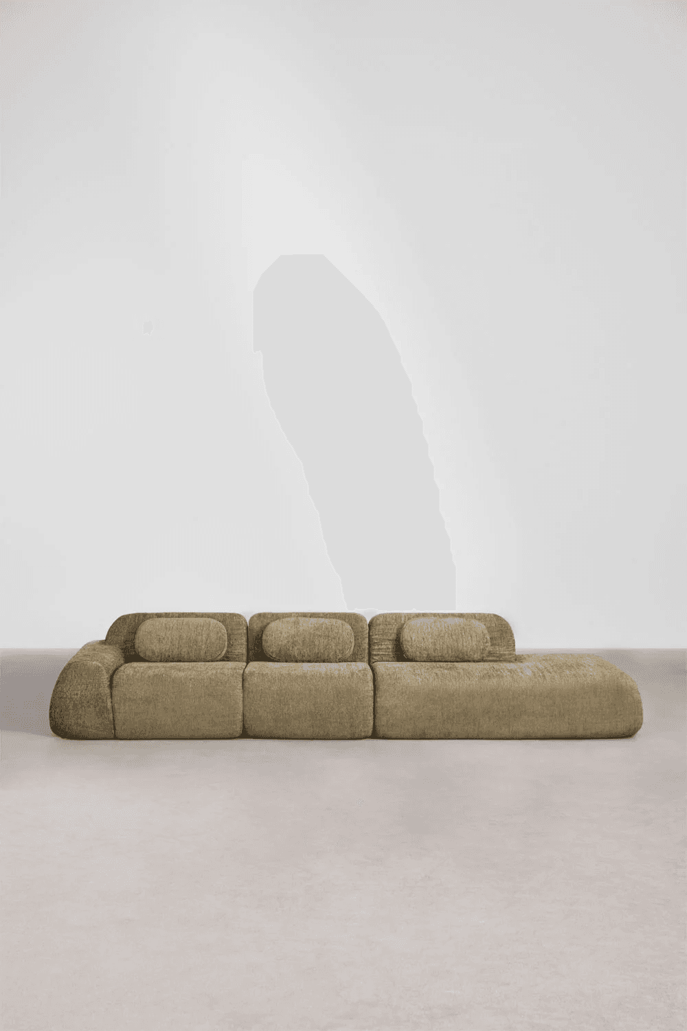 A low-profile contemporary sectional sofa with a chaise extension, upholstered in a textured, possibly bouclé or chenille fabric in a muted olive green or beige tone. The sofa is composed of three distinct modular sections: two armless sofa segments and one chaise lounge segment. Each segment features soft, rounded edges and integrated back cushions that are visually distinct but part of the continuous upholstered form, creating a plush and inviting aesthetic. — 3D model