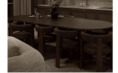 A modern dining set consisting of an oval table and six matching chairs, all in a dark brown finish. The table features a substantial oval top and a sculptural, broad base with geometric cutouts. The chairs are characterized by their rounded, continuous backrests and integrated armrests, upholstered in a dark brown fabric that matches the table's finish. The chairs are designed to fit closely around the table. — 3D model
