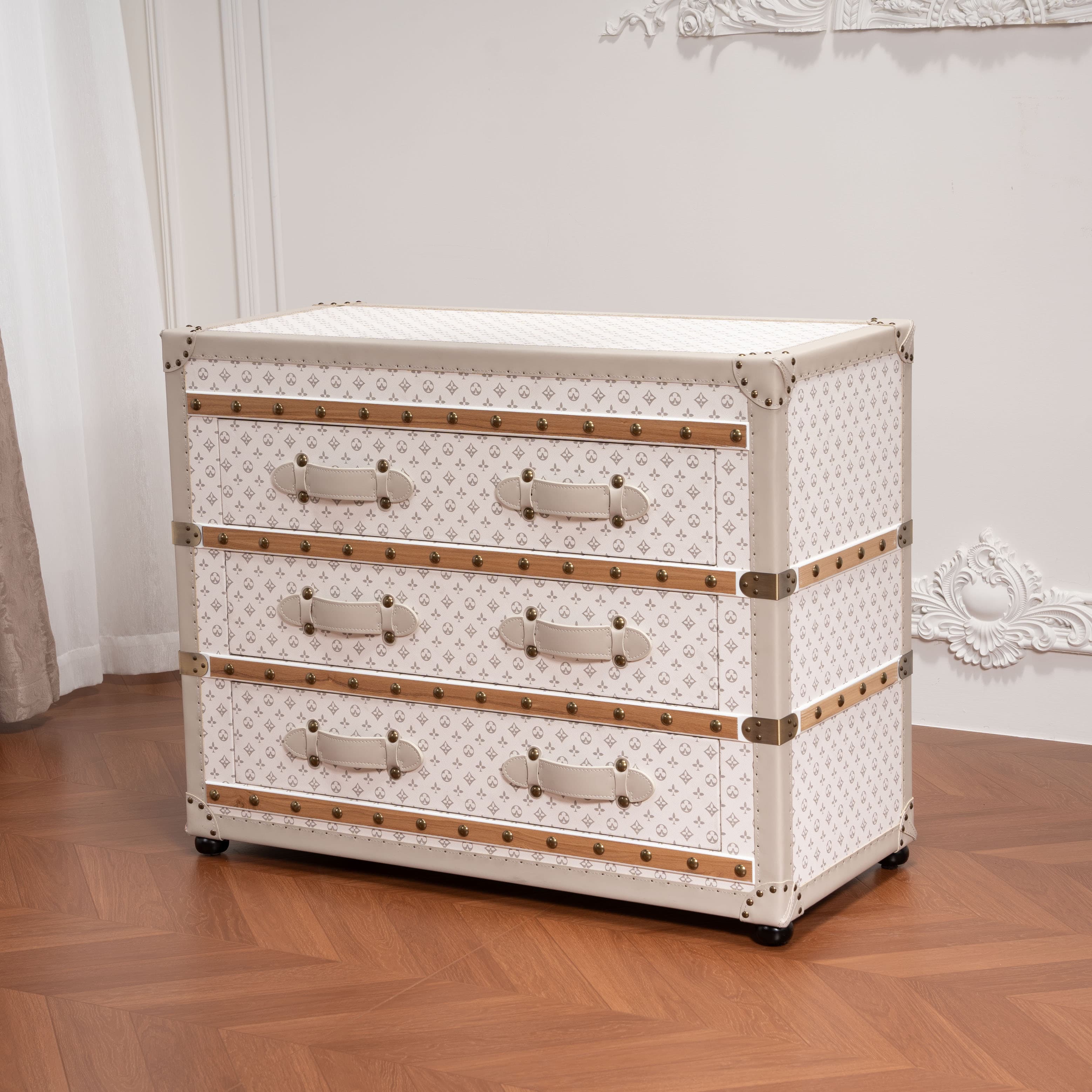 A chest of drawers designed to resemble a vintage travel trunk. It features three drawers, each with leather-like pull handles and decorative brass-colored hardware. The exterior is covered in a light-colored material with a repeating pattern, accented with beige trim and metal corner protectors. The piece is elevated by small black wheels at the base, suggesting mobility. — 3D model