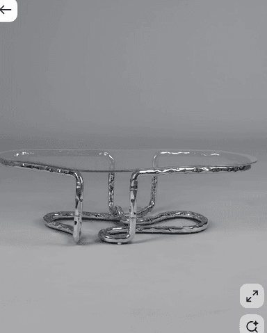 A low-profile coffee table featuring an irregularly shaped clear glass top supported by a sculptural base made of hammered silver-toned metal tubing bent into an abstract, organic form. — 3D model