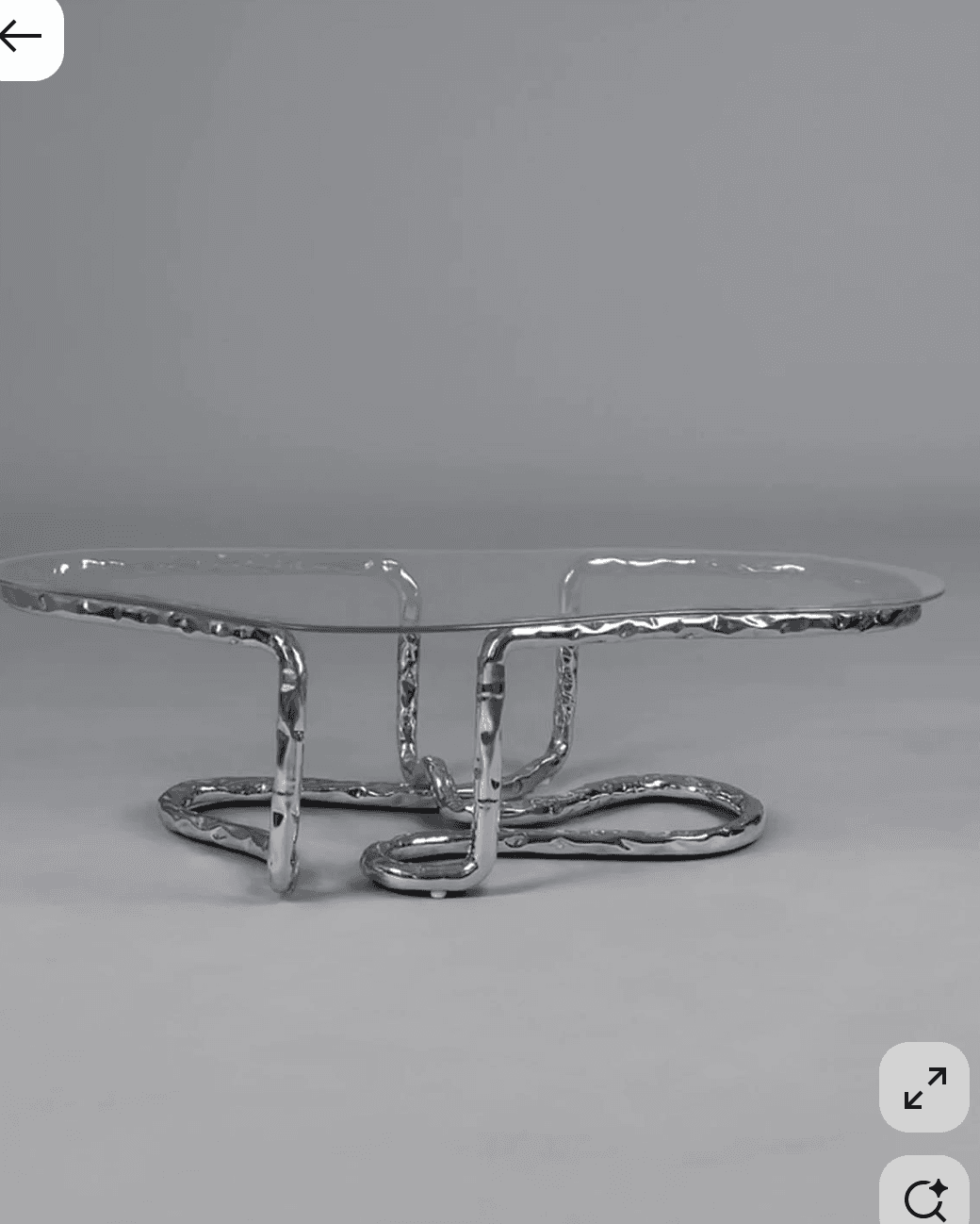 A low-profile coffee table featuring an irregularly shaped clear glass top supported by a sculptural base made of hammered silver-toned metal tubing bent into an abstract, organic form. — 3D model