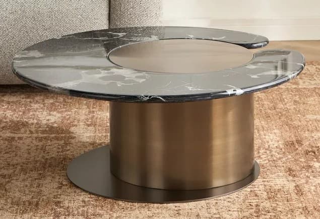 A contemporary coffee table featuring an irregularly shaped black marble top with white veining. The tabletop includes a central circular brushed metallic insert designed to swivel. The entire top is supported by a large, brushed brass cylindrical base, which in turn rests on a flat, circular black metal plate. — 3D model