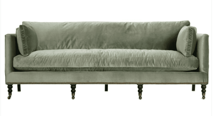 A traditional three-seater sofa upholstered in a light sage green velvet. It features classic English roll arms, loose seat and back cushions, and two round bolster pillows. The base of the sofa is adorned with antique brass nailhead trim, and it stands on five turned dark wooden legs, with the two end legs having caster wheels. — 3D model