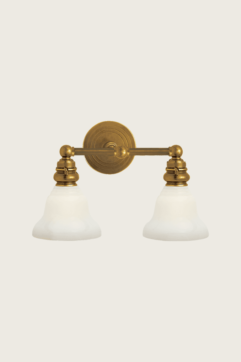 The image displays a traditional two-light wall sconce featuring an aged brass finish and frosted bell-shaped glass shades. The fixture consists of a circular backplate with concentric rings, from which a horizontal arm extends. Two lamp holders, each adorned with decorative brass elements, are attached to this arm, suspending the downward-facing glass shades. The overall design suggests a classic or transitional aesthetic. — 3D model