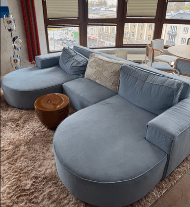 A modular sectional sofa with a light blue upholstered finish. The sofa features a curved chaise lounge section on the left and a straight section with an armrest on the right. It includes several back cushions and one decorative throw pillow with a textured pattern. The overall design is contemporary and low-profile. — 3D model