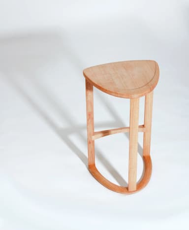 A wooden bar stool with a triangular, slightly dished seat and three cylindrical legs. One leg is positioned at the front and two at the back. A curved stretcher connects the two back legs near the bottom, and a diagonal stretcher connects the front leg to the rear left leg. The stool is constructed from light-colored wood with a visible grain. — 3D model