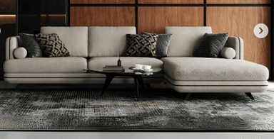 An L-shaped sectional sofa featuring a right-hand facing chaise lounge, upholstered in a light, neutral-toned fabric. The design is contemporary, characterized by clean lines, rounded edges on the arms and back, and distinctive dark contrast piping tracing the seams along the base, arms, and back cushions. The sofa sits on short, dark, tapered legs. It is adorned with several patterned throw pillows in contrasting dark grey and brown tones, adding visual interest to the minimalist base. — 3D model