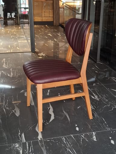 A modern dining chair constructed from a light brown wooden frame, featuring a distinctive rounded solid wood backrest and a channel-tufted upholstered seat in a reddish-brown (maroon) faux leather material. The chair's design emphasizes clean lines and a contemporary aesthetic, with the backrest appearing to be integrated seamlessly into the rear leg supports. — 3D model