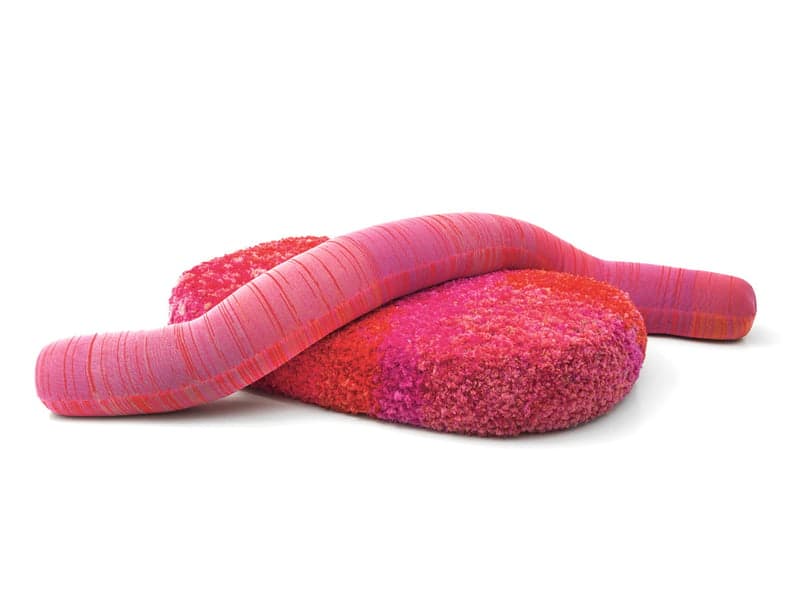 An organically shaped ottoman, possibly part of a modular seating system. It features a rounded, cushion-like base upholstered in a dense, shaggy fabric. A long, tubular element, upholstered in a fabric with a fine ribbed texture, is draped over the base, creating a dynamic, sculptural form. The color palette consists of various shades of pink and red, with the tubular element exhibiting a gradient effect. — 3D model