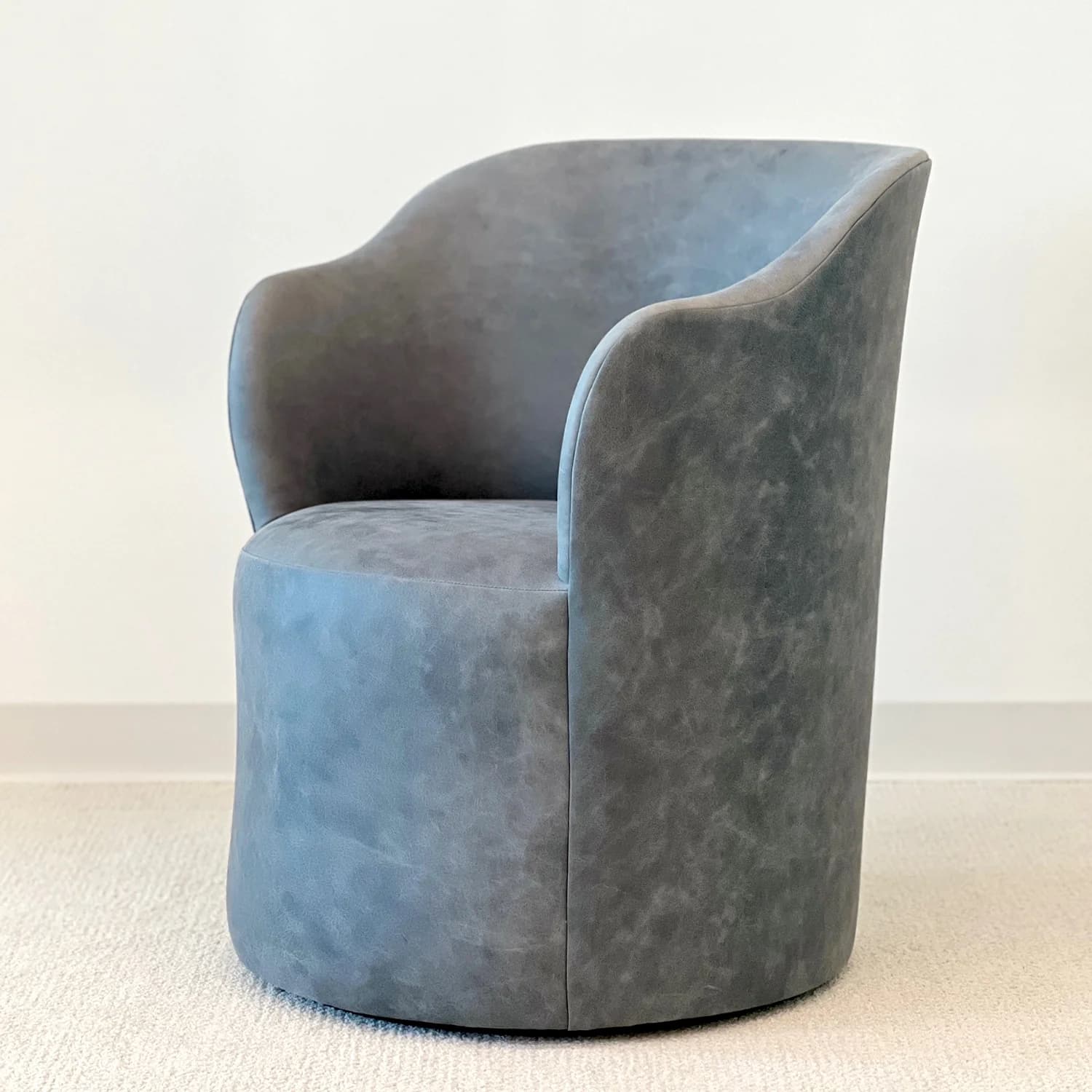A contemporary armchair with a rounded, barrel-like base and a gracefully curved backrest that extends into arms. The upholstery has a textured, somewhat distressed appearance, suggesting a soft fabric or faux leather. The chair is presented against a neutral background, resting on a textured carpet. — 3D model