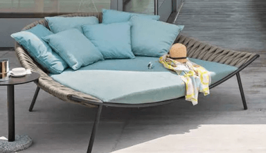 A modern outdoor daybed featuring a semi-circular form with a tightly woven gray synthetic rope backrest and dark metal legs. It is fitted with a large, light blue primary cushion and several smaller matching throw pillows. A straw hat and a patterned towel are placed on the cushion, suggesting a leisure context. — 3D model