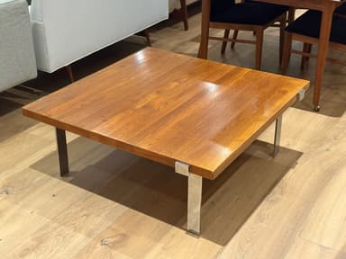 A square coffee table featuring a wooden top and four recessed metal legs. The tabletop has a polished, reddish-brown finish, suggesting a teak or walnut construction. The legs are made of polished chrome or stainless steel, with a rectangular profile, and are integrated into the corners of the table, giving it a minimalist and sleek appearance. The table exhibits characteristics of Mid-Century Modern design. — 3D model