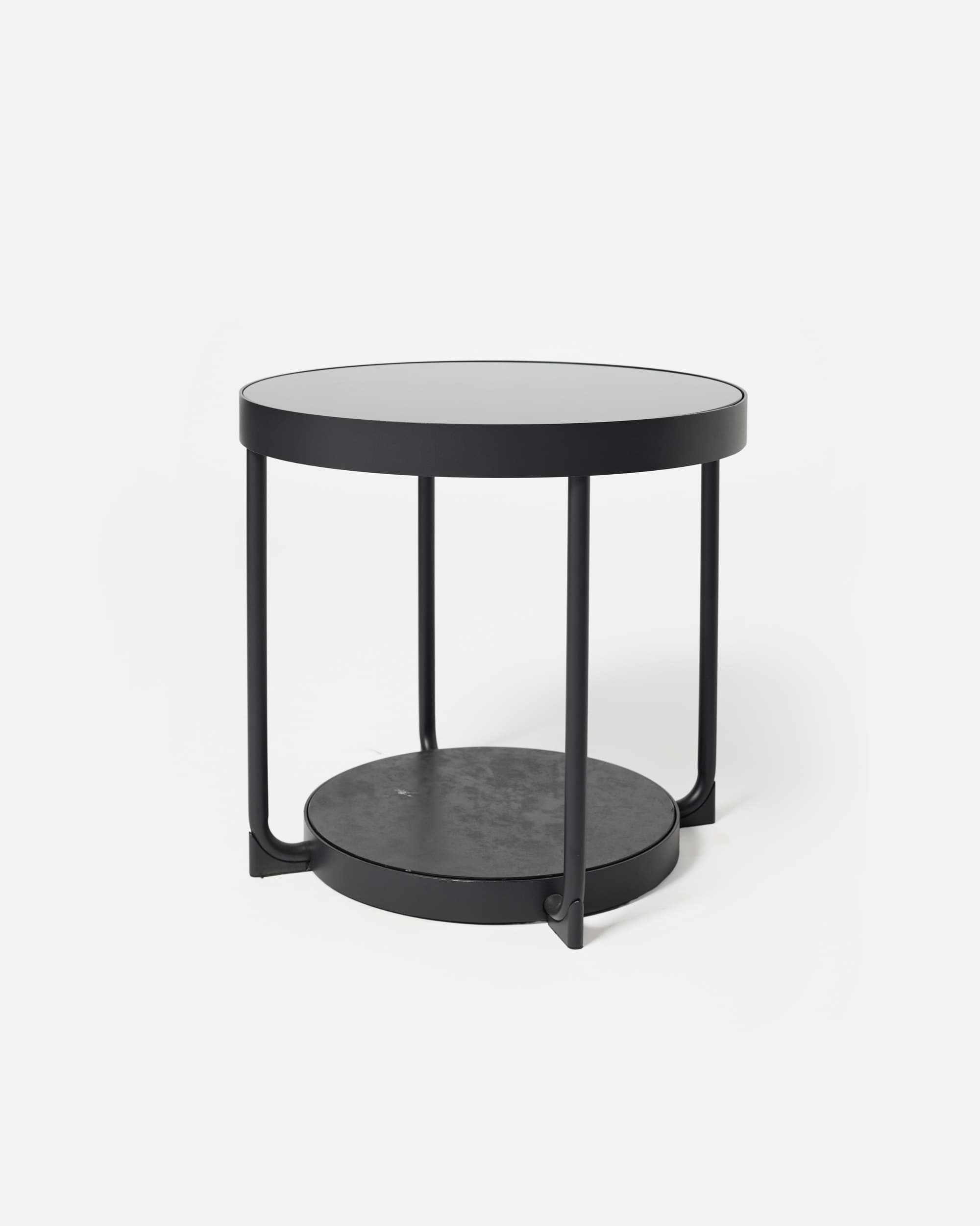 A modern, minimalist round side table featuring two circular tiers. The table is constructed from black-coated metal, with a smooth upper surface and a slightly textured lower shelf. Four cylindrical legs support the top tier, extending downwards to form the base structure that holds the lower shelf. The overall design is sleek and functional. — 3D model