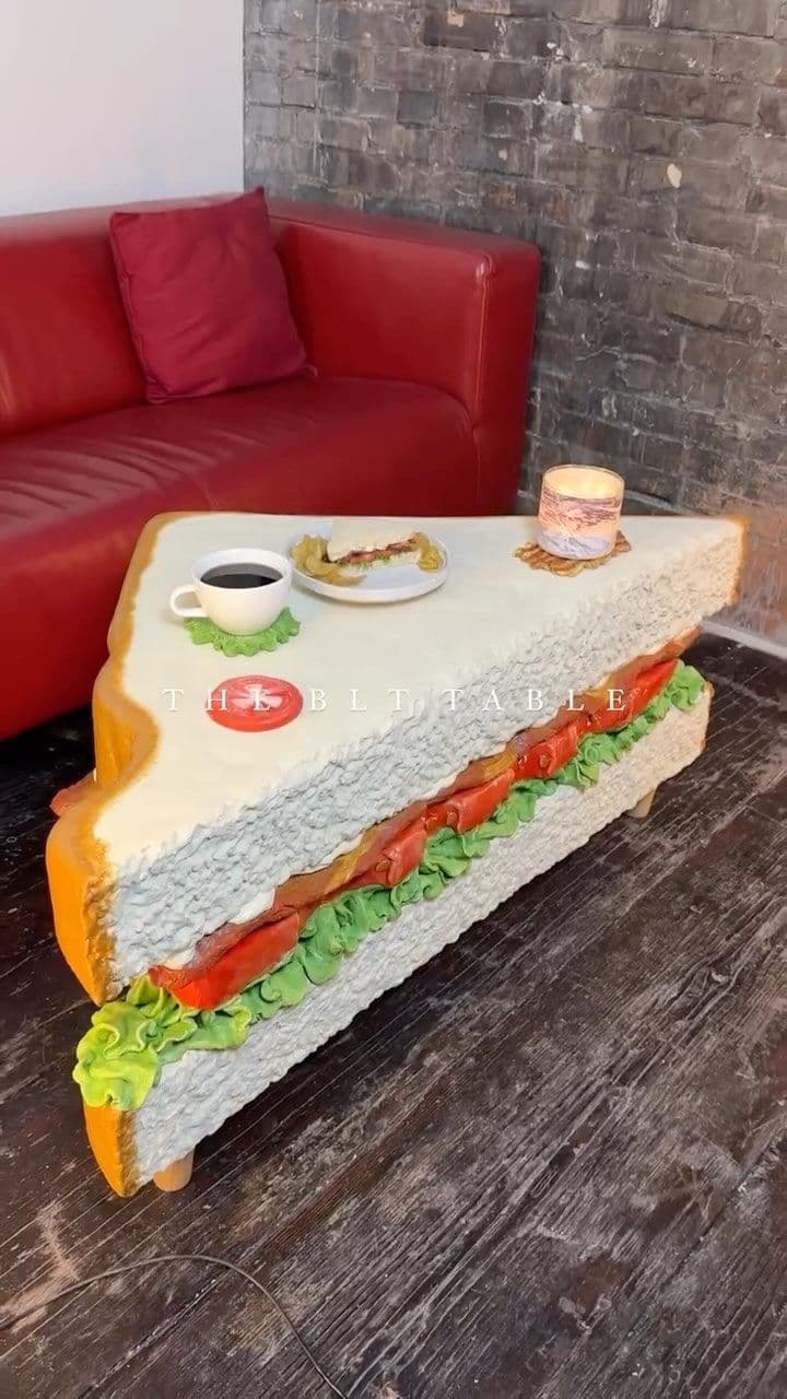 This image features a novel and whimsical accent table designed to resemble a large, diagonally-cut club sandwich. The table's surface is the 'top slice' of bread, with a visible crust edge. Below this, layers mimicking lettuce (green ruffled texture), tomato slices (red ovals), and bacon strips (brown textured elements) form the sides of the table. Four short, light-colored cylindrical legs support the structure from the bottom 'slice' of bread. A miniature coffee cup on a green coaster and a small plate with a tiny sandwich rest on the table's surface, indicating its scale and function. — 3D model