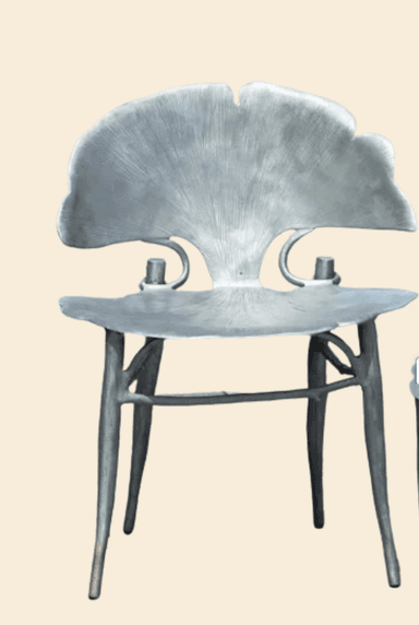 A sculptural dining chair crafted from cast metal, featuring an organic design reminiscent of a ginkgo leaf. Both the backrest and seat are shaped to mimic the characteristic fan-like form, complete with a textured, ribbed surface that suggests natural veining. The chair is finished in a uniform silver-grey tone, indicating a brushed or sandblasted metallic surface. It stands on four slender, tapered legs that seamlessly integrate into the underside of the seat, creating a cohesive and artful silhouette suitable for contemporary interiors. — 3D model