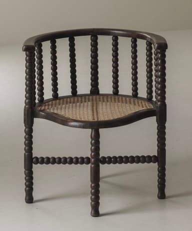 A dark stained wooden barrel armchair featuring a bobbin-turned frame and a woven cane seat. The back and armrests form a continuous curved structure, supported by vertical bobbin-turned spindles. The four bobbin-turned legs are connected by bobbin-turned stretchers forming an X-shape beneath the seat. The seat itself is crafted from woven cane with a slightly curved front. — 3D model