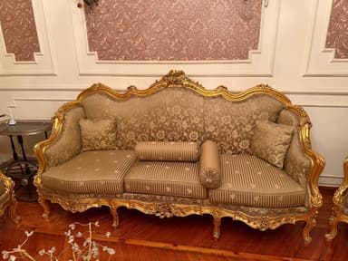 An opulent three-seater sofa characterized by an elaborate, hand-carved wooden frame finished in gold leaf. The sofa features a curvilinear backrest, armrests, and apron adorned with intricate scrollwork. It is upholstered in a rich, patterned beige and gold fabric, with striped seat cushions. The piece rests on decorative cabriole legs and includes two square throw pillows and two cylindrical bolster pillows. — 3D model