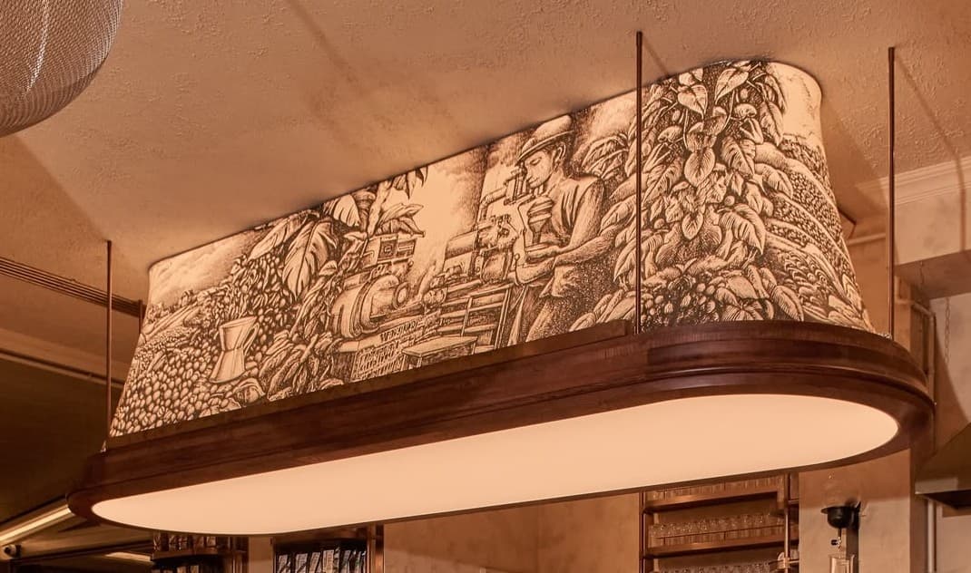 A rectangular, curved pendant light featuring a dark wood frame and an intricately illustrated outer shade. The shade depicts a narrative scene, possibly related to coffee cultivation, in a sepia-toned, etched style. Below the illustrated shade is a frosted, translucent diffuser that emits a soft, warm light. The fixture is suspended by four thin rods or cables, suggesting it is designed to hang over a surface. — 3D model