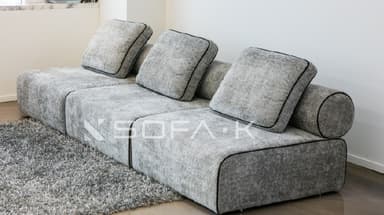 A contemporary, low-profile modular sofa upholstered in a textured, speckled grey fabric. The sofa consists of three distinct seating modules, each with a loose seat cushion and a loose rectangular back cushion. The back cushions feature black piping around their edges. The sofa integrates low, rounded or rolled armrests at each end, which blend seamlessly with the overall low-profile design. A dark line of piping or stitching runs along the seams of the modular units and along the bottom edge of the sofa, creating a clean, defined aesthetic. The sofa rests directly on the floor, lacking visible legs. — 3D model