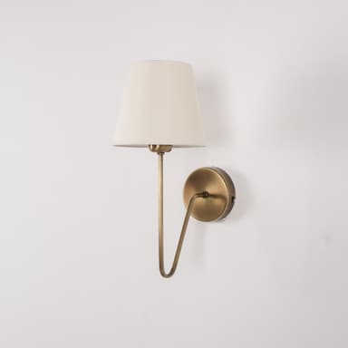 A wall-mounted lamp with a conical, cream-colored fabric shade and a brass-finished metal arm and base. The arm has a gentle curve and extends from a circular base plate mounted on the wall. The shade is open at the bottom and tapers upwards. — 3D model