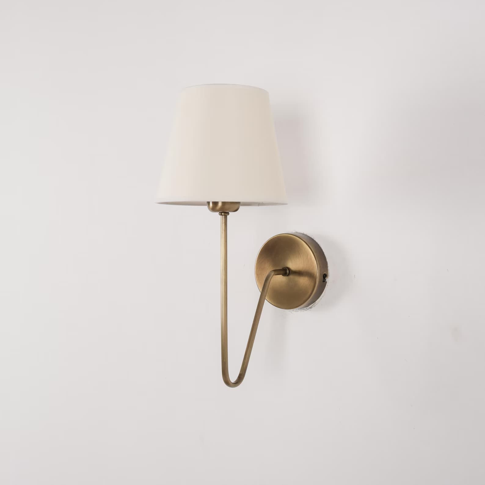 A wall-mounted lamp with a conical, cream-colored fabric shade and a brass-finished metal arm and base. The arm has a gentle curve and extends from a circular base plate mounted on the wall. The shade is open at the bottom and tapers upwards. — 3D model