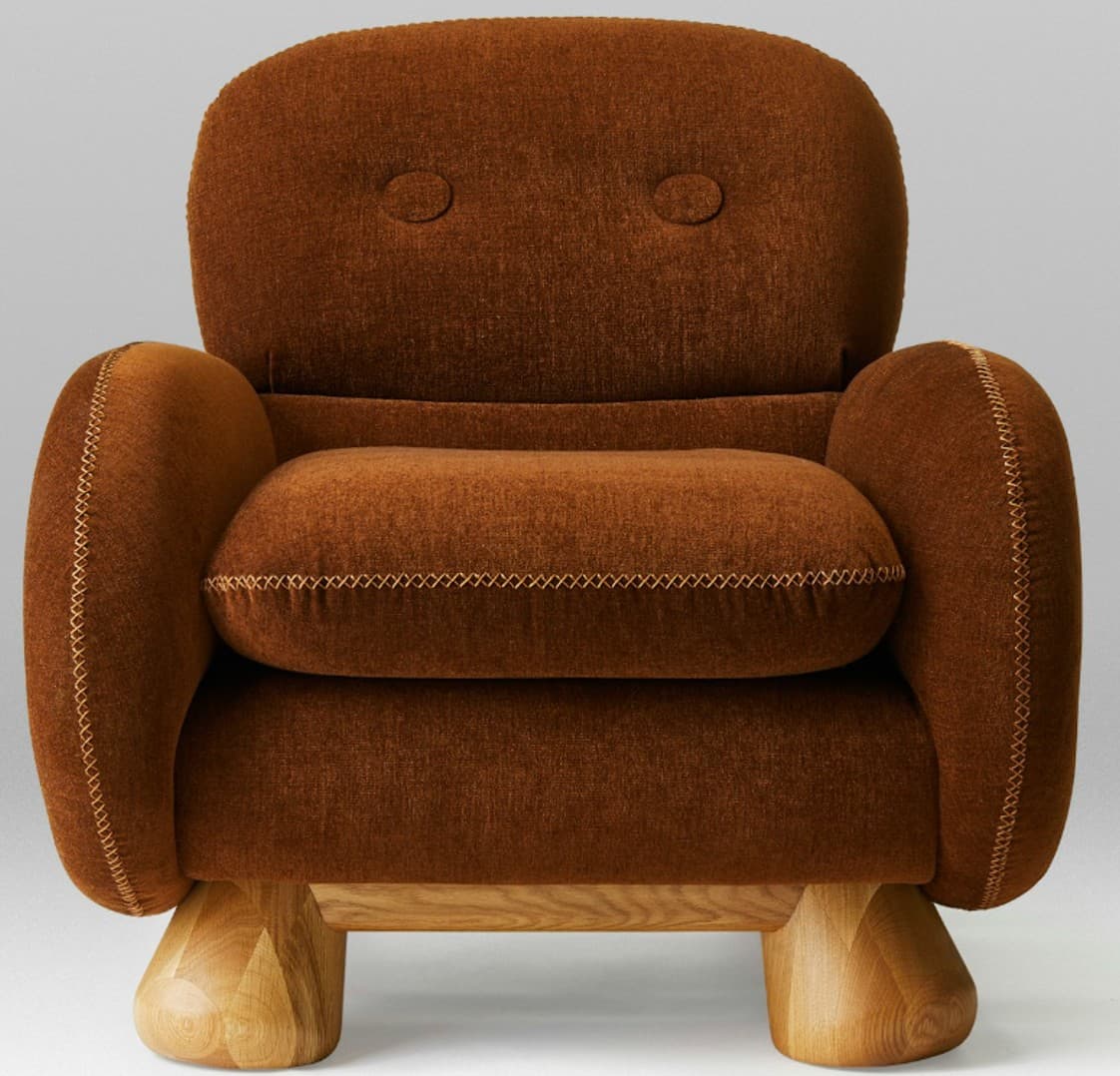 A generously sized armchair upholstered in a textured, deep brown fabric. The chair features rounded arms and a high back with two circular tufts resembling eyes. Prominent stitching in a contrasting light brown thread outlines the arms, seat cushion, and back, giving it a handcrafted aesthetic. The seat cushion is thick and appears overstuffed. The chair is supported by four distinct, rounded wooden legs with faceted detailing near the base, showcasing a natural wood grain. — 3D model