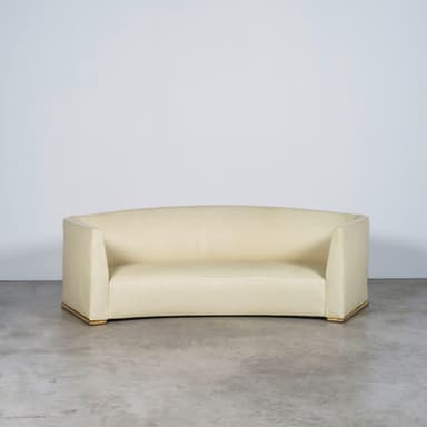A contemporary curved sofa upholstered in light beige fabric, featuring a continuous back and armrest design, and a recessed plinth base accented with a gold-toned trim. — 3D model