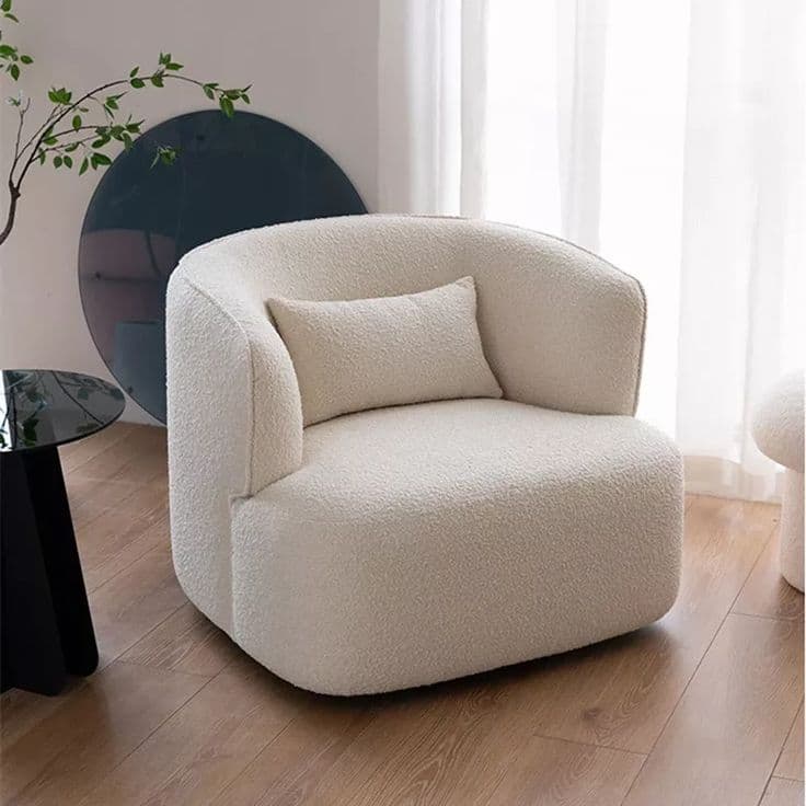 A contemporary, fully upholstered barrel-style lounge chair in a cream-colored boucle fabric. The chair features a rounded, continuous backrest that flows into integrated armrests, creating a cohesive and inviting silhouette. It has a deep, wide seat designed for comfort and is accompanied by a small, matching rectangular accent pillow. The chair sits low to the ground with a robust, unexposed base, maintaining its sleek and modern aesthetic. The boucle fabric provides a textured, soft appearance. — 3D model
