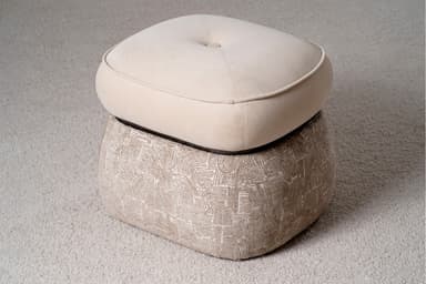 A two-tiered ottoman with a rounded, tufted top cushion and a patterned base. The top cushion is upholstered in a smooth, light beige fabric and features a single button tuft in the center, outlined by stitched seams. A contrasting piping trim defines the edge of the top cushion. The base is wider than the top and is upholstered in a textured fabric with a repeating abstract pattern in shades of beige and white, resembling arcs and linear shapes. The ottoman is situated on a textured, light-colored carpet. — 3D model