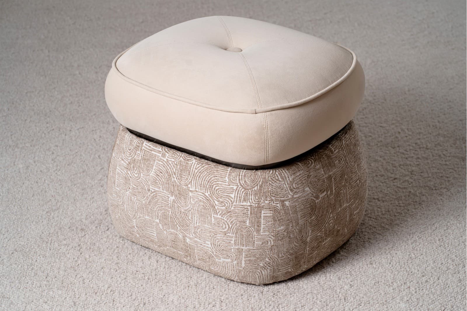 A two-tiered ottoman with a rounded, tufted top cushion and a patterned base. The top cushion is upholstered in a smooth, light beige fabric and features a single button tuft in the center, outlined by stitched seams. A contrasting piping trim defines the edge of the top cushion. The base is wider than the top and is upholstered in a textured fabric with a repeating abstract pattern in shades of beige and white, resembling arcs and linear shapes. The ottoman is situated on a textured, light-colored carpet. — 3D model