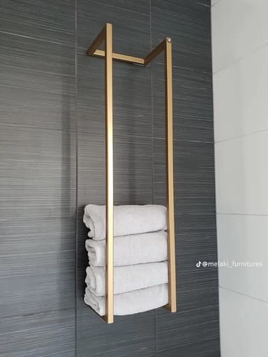 A minimalist, rectangular, floor-standing towel stand constructed from gold-colored metal. It features a simple, open frame design, allowing for the vertical storage of stacked towels. Four light grey, folded towels are visible, resting within the lower section of the frame. The background is plain white, emphasizing the sleek design of the stand and its contents. — 3D model