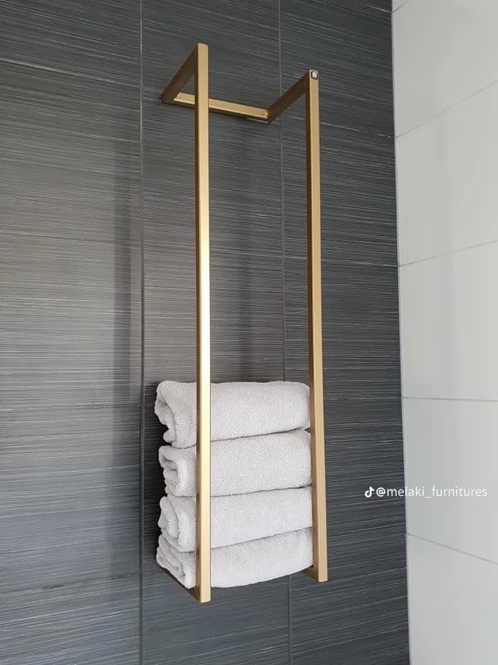A minimalist, rectangular, floor-standing towel stand constructed from gold-colored metal. It features a simple, open frame design, allowing for the vertical storage of stacked towels. Four light grey, folded towels are visible, resting within the lower section of the frame. The background is plain white, emphasizing the sleek design of the stand and its contents. — 3D model