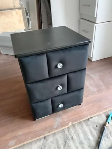 A sleek, black three-drawer storage unit, likely serving as a nightstand or small chest. It features a rectangular top and base, with three vertically stacked drawers. The drawer fronts are upholstered with a soft, velvet-like black fabric, each adorned with a prominent, clear faceted crystal-style knob. The body of the unit appears to be constructed from wood or MDF with a smooth, matte black finish. — 3D model