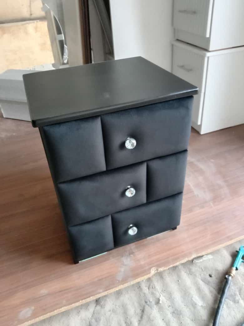 A sleek, black three-drawer storage unit, likely serving as a nightstand or small chest. It features a rectangular top and base, with three vertically stacked drawers. The drawer fronts are upholstered with a soft, velvet-like black fabric, each adorned with a prominent, clear faceted crystal-style knob. The body of the unit appears to be constructed from wood or MDF with a smooth, matte black finish. — 3D model