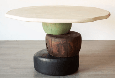 An accent table featuring an irregularly shaped, off-white plaster or concrete top, supported by a sculptural base composed of three stacked, cylindrical wooden blocks in varying colors and textures. The base consists of a light green fluted block, a dark brown natural wood block, and a black-stained, textured wood block at the bottom. — 3D model