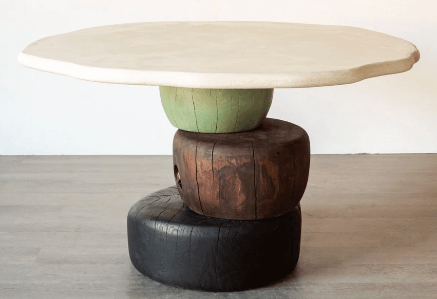 An accent table featuring an irregularly shaped, off-white plaster or concrete top, supported by a sculptural base composed of three stacked, cylindrical wooden blocks in varying colors and textures. The base consists of a light green fluted block, a dark brown natural wood block, and a black-stained, textured wood block at the bottom. — 3D model