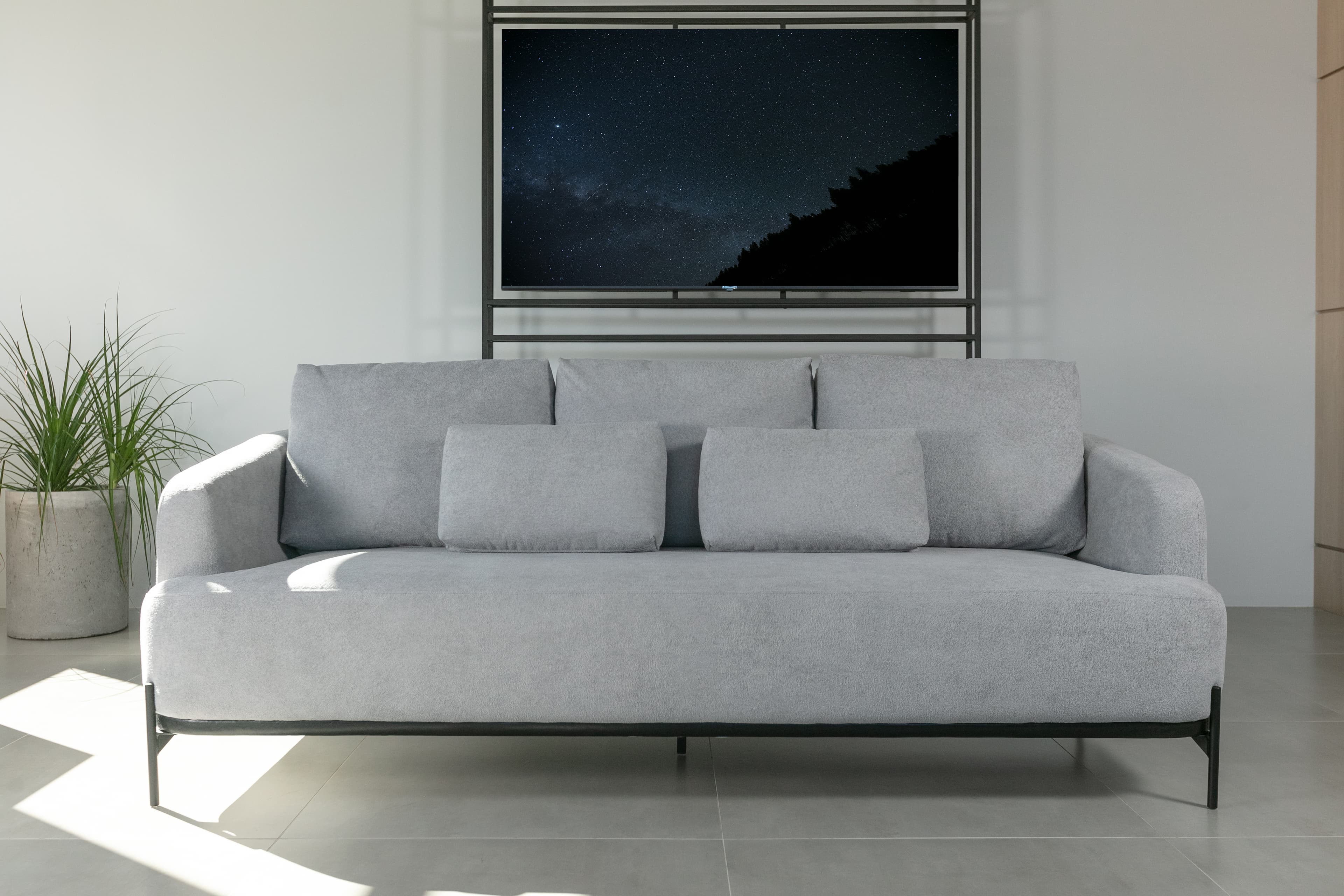 A contemporary three-seater sofa featuring light grey fabric upholstery. The sofa has a clean, minimalist design with subtly rounded arms and a low profile. It rests on a slender black metal frame that forms both the base and integrated legs, giving it an elevated appearance. The seating consists of a single large cushion, while the backrest is composed of three loose back cushions and two smaller rectangular throw pillows, all upholstered in matching light grey fabric. — 3D model