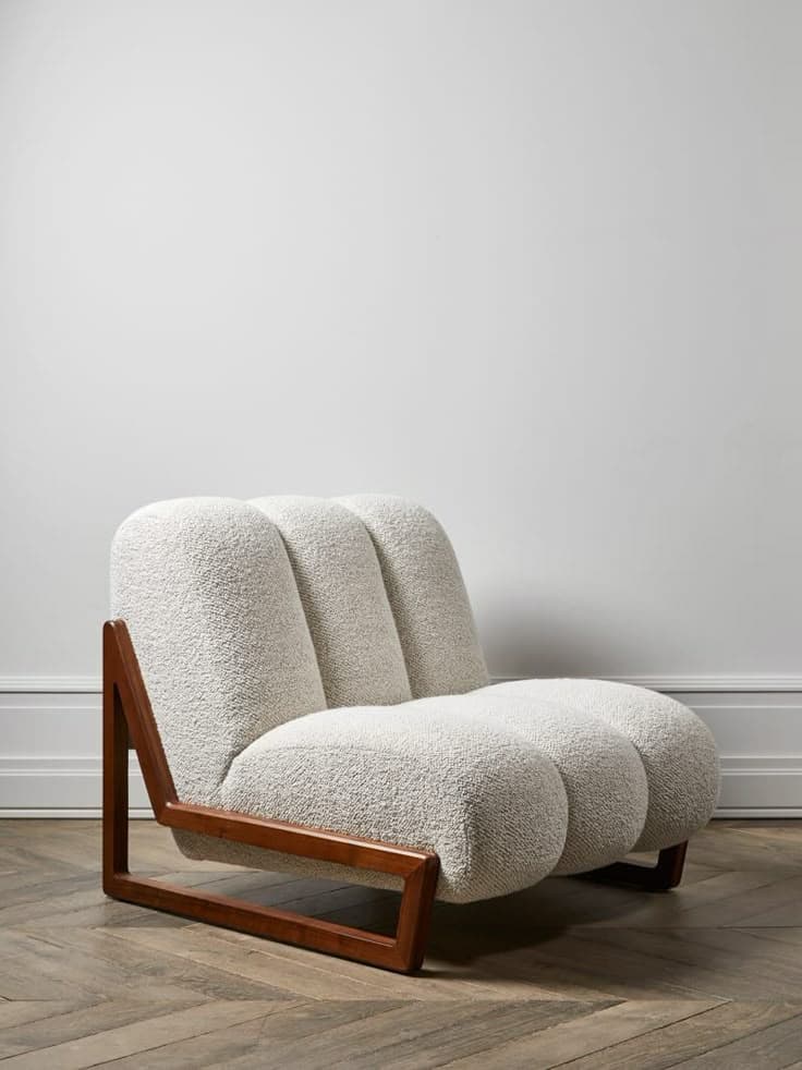 A modern armchair with a distinctive design. The seat and backrest are upholstered in a textured, boucle-like fabric and feature a series of rounded, segmented cushions. The frame is constructed from dark wood, exhibiting a geometric, angular shape on the left side, which contrasts with the softness of the upholstery. — 3D model
