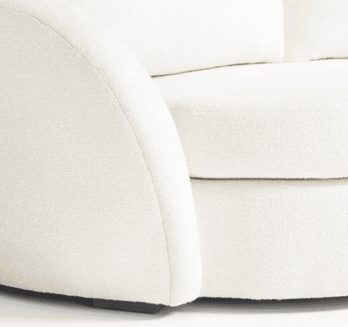 A close-up view of a cream-colored sofa, focusing on the armrest and seat cushions. The upholstery appears to be a textured boucle or similar woven fabric. The armrest has a rounded, curved profile, and the seat cushions are plush with visible stitching. The base of the sofa, where it meets the floor, is partially visible and appears to be a dark wood. — 3D model