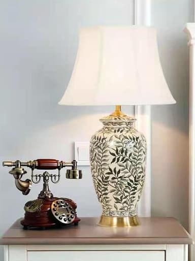 A decorative table lamp featuring an urn-shaped ceramic base adorned with a botanical leaf pattern in shades of gray and black. It sits on a gold-toned metal pedestal and is topped with a white tapered drum fabric shade. — 3D model