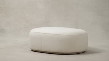 A contemporary upholstered ottoman with a rounded oval shape, covered in a plush cream bouclé fabric. The design features soft, curvilinear forms and a low-profile base, contributing to its modern and inviting aesthetic. A subtle seam runs along the perimeter of the top surface, connecting to vertical seams on the sides. — 3D model