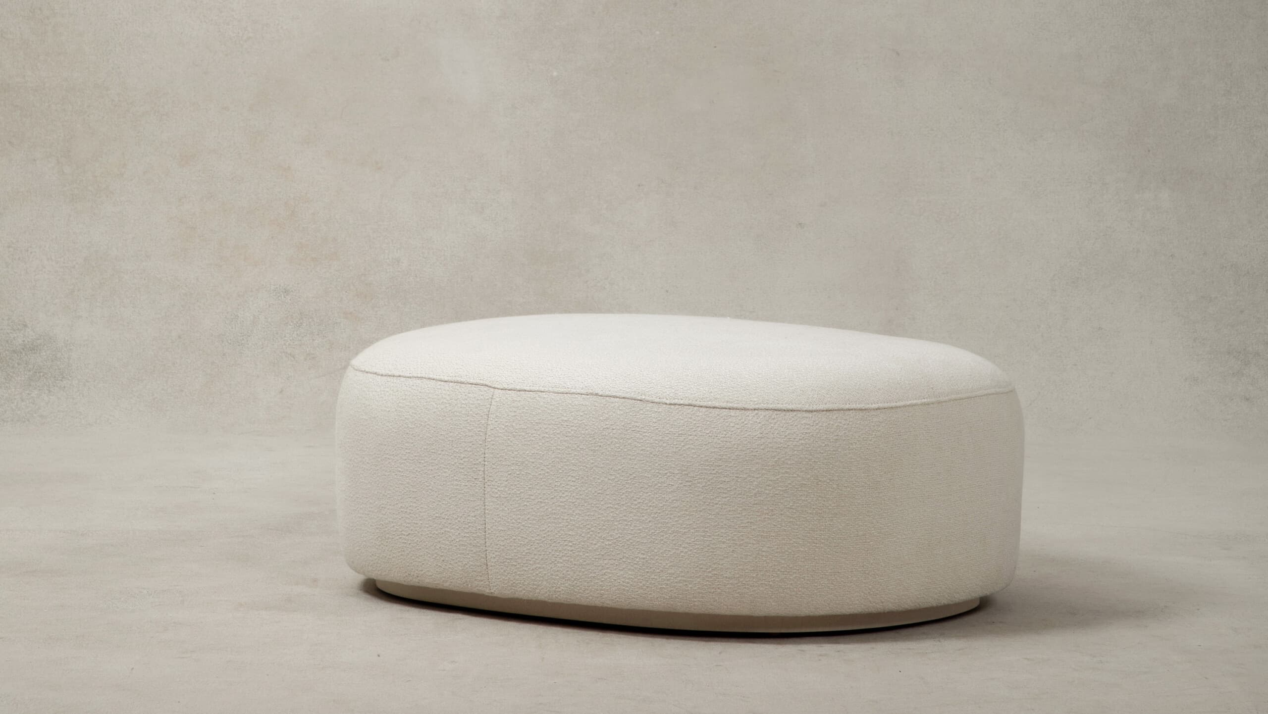 A contemporary upholstered ottoman with a rounded oval shape, covered in a plush cream bouclé fabric. The design features soft, curvilinear forms and a low-profile base, contributing to its modern and inviting aesthetic. A subtle seam runs along the perimeter of the top surface, connecting to vertical seams on the sides. — 3D model