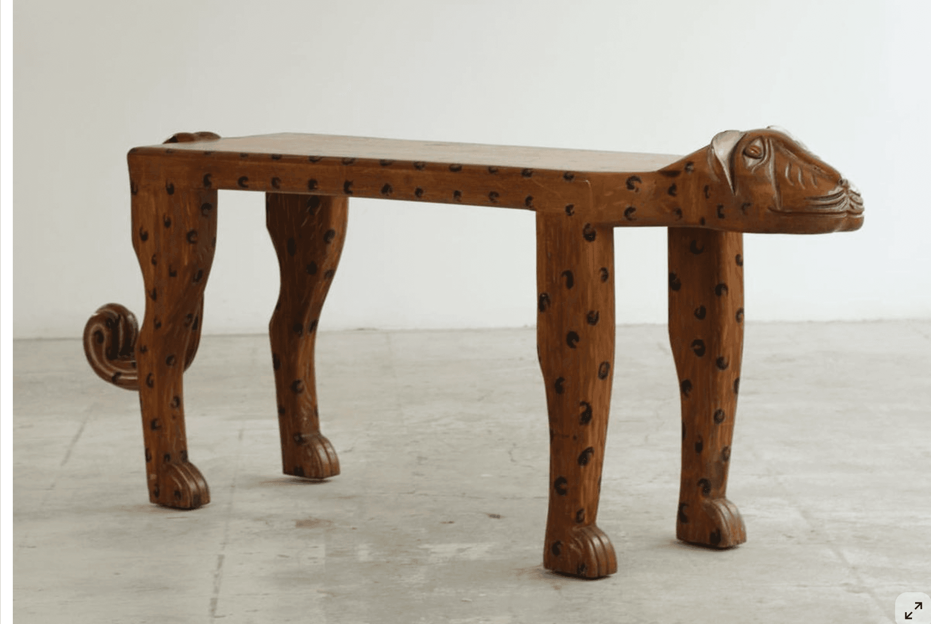 A wooden bench designed to resemble a cheetah. The seat is flat and rectangular, supported by four legs shaped like the animal's limbs. The head of the cheetah is carved at one end of the bench, and a tail is curled at the other. The surface of the bench and the legs are decorated with black spots, mimicking the coat of a cheetah. The overall aesthetic suggests an artistic or decorative piece. — 3D model