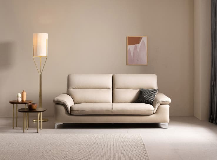 A light beige leather sofa with two seat cushions and a tufted seat base. It features a contemporary design with slightly sloped arms and a single row of tufting across the seat. The sofa is supported by slender metal legs. A single dark grey throw pillow with a textured pattern rests on the right seat cushion. — 3D model