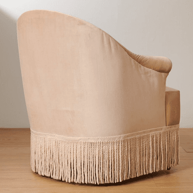A light pink velvet armchair with a rounded back and arms. It features decorative pleating on the armrests and a prominent fringe along the bottom base. The seat cushion is a lighter, textured fabric. The chair has short, dark wooden legs visible at the very bottom. — 3D model